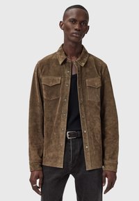 Bruine suède overhemdjack met twee borstzakken, drukknoopsluiting en een puntkraag, gedragen over een zwart shirt en donkere jeans.
