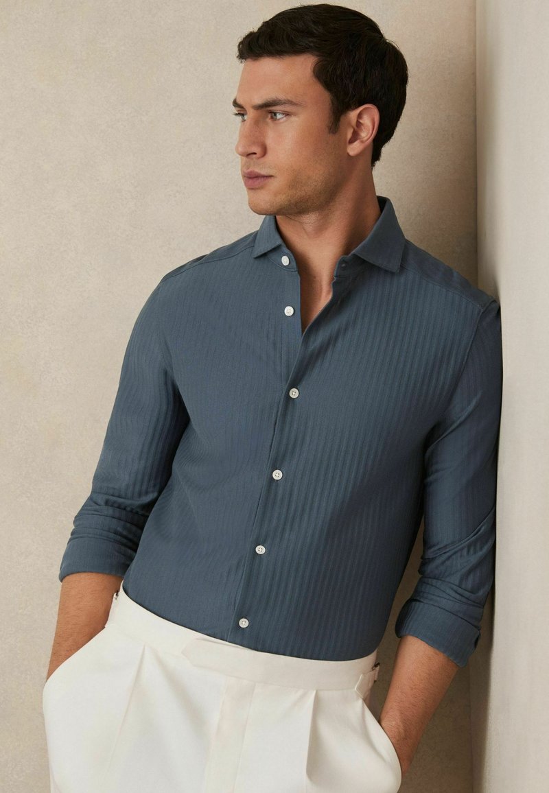 Camisa de rayas azul oscuro con cuello, mangas largas arremangadas y botones blancos. Se combina con pantalones ajustados de color crema.