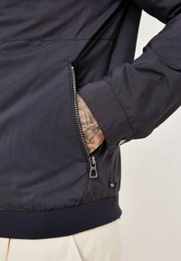 Dark navy zippered jacket with vertical stripes, ribbed cuffs, and hem; features a side pocket with a metallic zipper. Textured fabric.