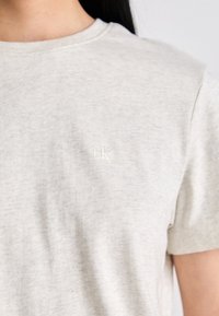 Svetlošedé tričko s krátkym rukávom a malým bielym vyšitým logom "cK" na hrudi, nosené osobou s dlhými tmavými vlasmi.