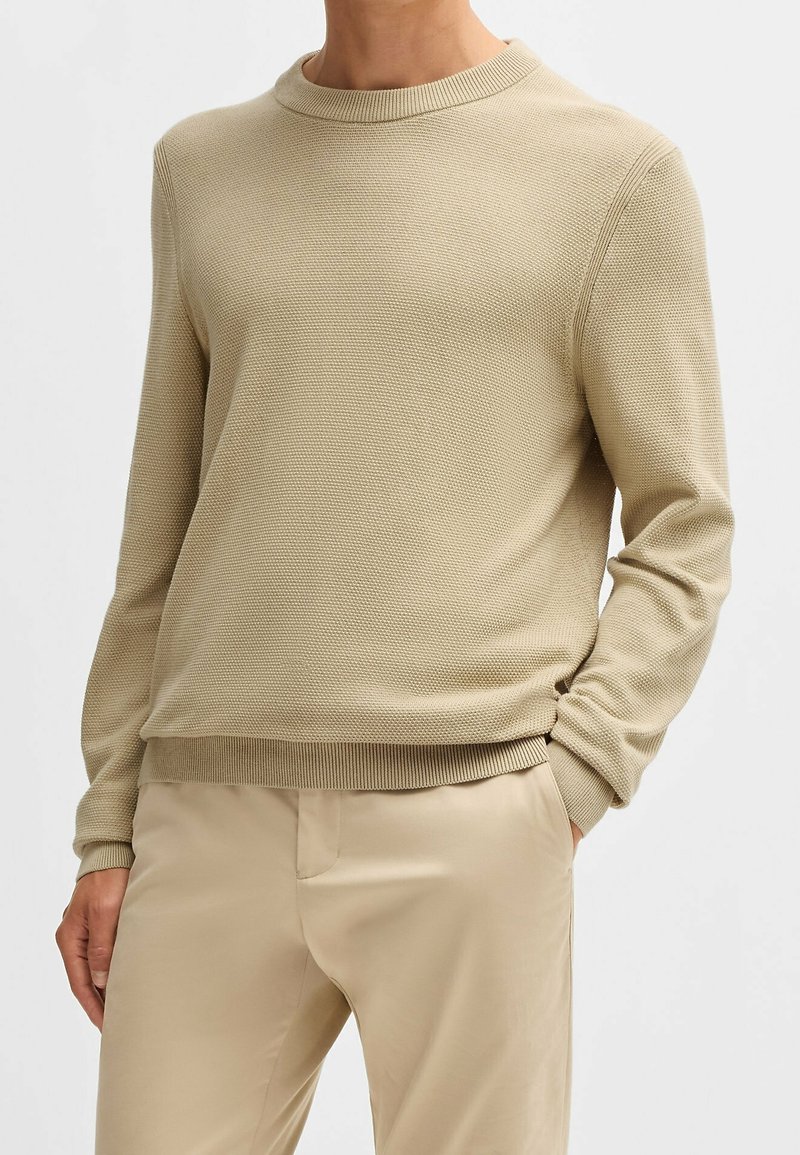 Homme portant un pull en maille beige texturée et un pantalon beige assorti, avec une main dans la poche, sur un fond blanc uni.