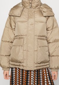 Beige polstret jakke med høj krave, trykknapklapper og frontlommer. Stoffet har en glat tekstur med en blank overflade.