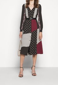 Žena v páskových dlouhých rukávech, s patchwork midi šaty v černobílých a burgundských vzorech, drží béžovou kabelku a má na sobě černé lodičky.