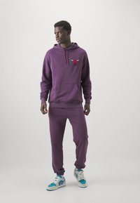 Sudadera morada con bolsillo frontal y logotipo bordado, combinada con pantalones joggers a juego y zapatillas coloridas con acentos en azul y aqua.