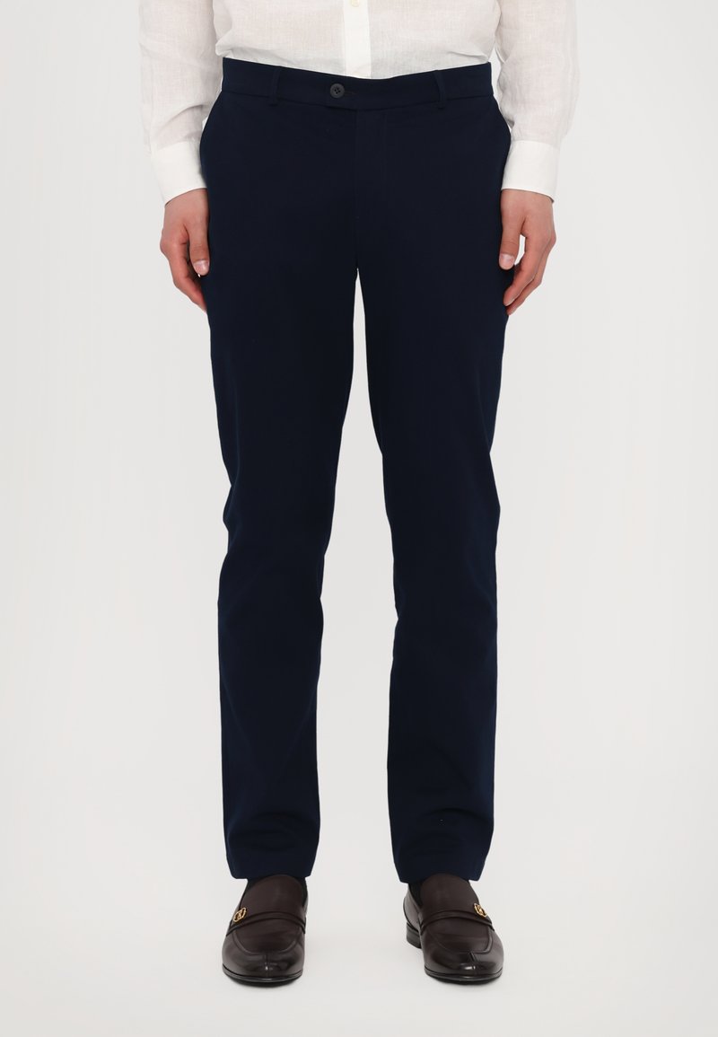 Sötétkék nadrág, szövetből készült, slim fit kialakítással, gombos záródással és oldalsó zsebekkel, barna mokaszinnal párosítva.