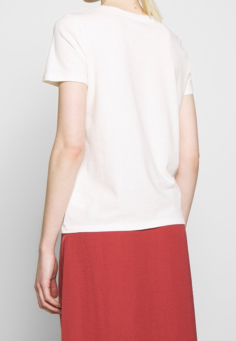 Kurzärmliges T-Shirt mit Rundhalsausschnitt aus leichtem cremefarbenem Baumwollstoff. Die Rückansicht zeigt eine lässige Passform. Kombiniert mit einem einfarbigen roten Rock, Rocklänge bis zur Mitte der Wade.