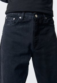 Vaqueros de mezclilla oscura con un diseño clásico de cinco bolsillos, botón metálico y textura sutil, que presentan un corte recto y un lavado oscuro.