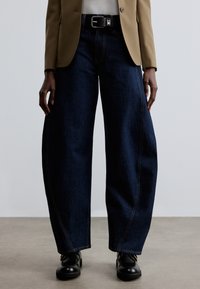 Laiade tumedad sinised denimpüksid nähtava õmblusega, kõrge vöökoht ja kontrastse musta vööga. Kombineeritud beežide bleiseri ja mustade kingadega.