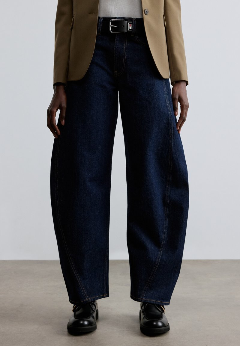 Tamsiai mėlynos spalvos platėjančios džinsinės kelnės su matomomis siūlėmis, aukštu liemeniu ir kontrastingu juodu diržu. Derinamos su smėlio spalvos švarku ir juodais batais.