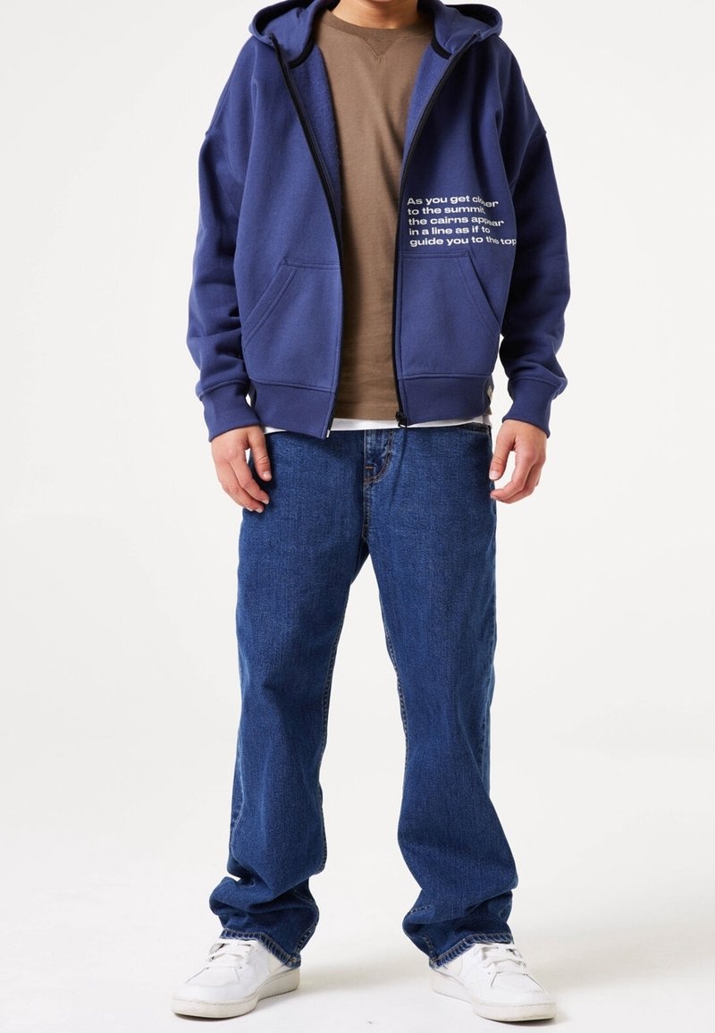 Homme portant un sweat à capuche bleu zippé avec texte, un t-shirt marron, un jean bleu et des baskets blanches, debout devant un fond blanc uni.