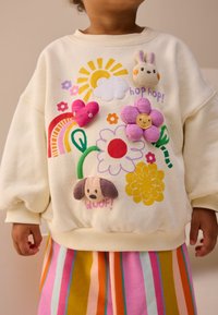Sweat-shirt couleur crème avec des motifs brodés colorés représentant des fleurs, des nuages, un lapin, un cœur et un chien. Associé à une jupe rayée.