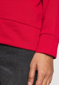 Felpa rossa in cotone con scollo a giro e polsini a costine, abbinata a jeans grigi scuri. Il tessuto appare liscio e leggermente strutturato.