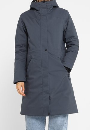 Kobieta w długim, ciemnoszarym zimowym płaszczu z kapturem, z przednimi kieszeniami i ukrytym zapięciem na guziki, nosząca jasnoniebieskie dżinsy.