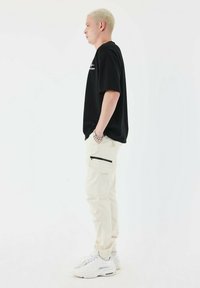 Sort t-shirt, beige cargo bukser med sidelommer og sorte lynlåse, og hvide sneakers. Håret er kort og lyst.