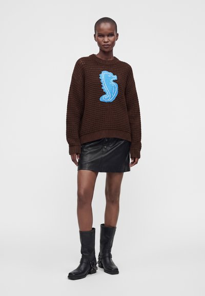 Femme portant un pull en maille marron avec un grand hippocampe bleu, une mini-jupe en cuir noire et des bottes noires mi-mollet, debout devant un fond uni.