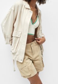 Veste beige oversize avec des boutons-pression, en tissu texturé et deux poches à l'avant, portée sur un crop top tricoté et un short cargo beige.