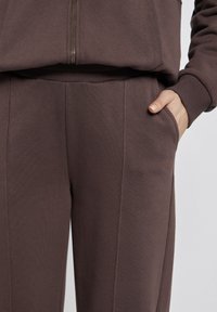 Brązowy komplet bluzy i spodni wykonany z miękkiego materiału, z ribbowymi ściągaczami, zapięciem na zamek oraz bocznymi kieszeniami dla dodatkowej wygody.