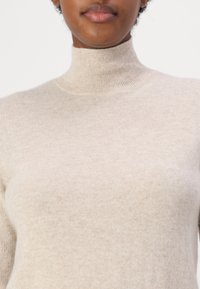 Ljusbeige tröja med hög krage gjord av mjukt, texturerat material. Har ribbad krage och subtila nyansvariationer.