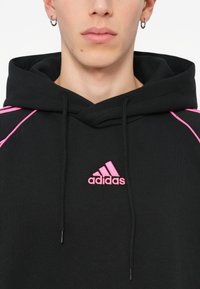 Čierna mikina s kapucňou a ružovými akcentmi, vybavená šnúrkou na stiahnutie a vyšitým logom Adidas na hrudi. Mäkká textúra a športový dizajn.