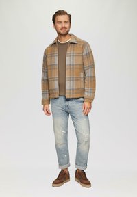 Bram zimski plašč v rjavi karo s sivo obrobo, v kombinaciji z rjavim puloverjem, svetlo modrimi raztrganimi jeans hlačami in rjavimi supergami.