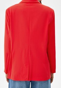 Punainen bleiseri, jossa on sileä, jäsennelty muotoilu, kaksilapainen kaulus, pitkät hihat ja yksittäinen takahalkio. Päällä sinisen denimin kanssa.