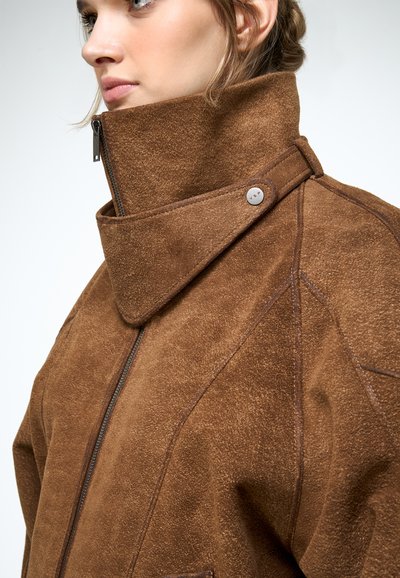 Žena v hnedej semišovej bunde s vysokým golierom zapnutým kovovým gombíkom a viditeľným predným zipsom.