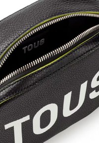 Bolsa de cuero negro con un acabado texturizado, presenta un logo blanco de "TOUS" y un ribete verde en la cremallera. El interior muestra un forro de marca.