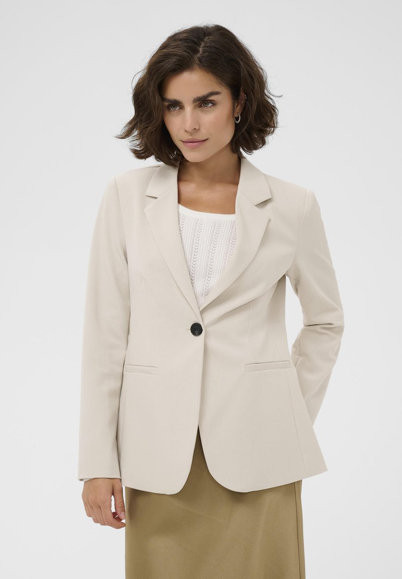 Femme aux cheveux courts et foncés portant un blazer beige sur un haut en maille blanche et une jupe kaki, debout devant un fond clair uni.