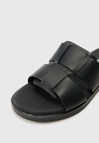 Crne kožne sandale na klizanje s širokim preklapajućim trakama i ravnim potplatom s glatkom završnom obradom. Minimalistički dizajn bez dodatnih ukrasa.