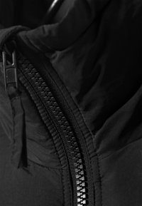 Schwarze Jacke mit Reißverschluss-Detail, strukturiertem Stoff und einem hohen Kragen. Der Reißverschluss ist verdeckt und fügt sich nahtlos in das Materialdesign ein.