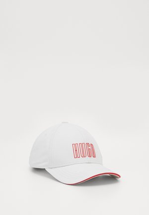 Weißes Baseball-Cap mit rotem "HUGO"-Logo auf der Vorderseite und rotem Rand entlang der Kante der gebogenen Krempe, auf einfarbigem Hintergrund.