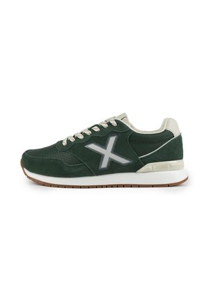 Zapatillas de ante verde con acentos beige, que presentan un logotipo plateado reflectante, detalles perforados y una suela blanca con una banda de rodadura marrón texturizada.
