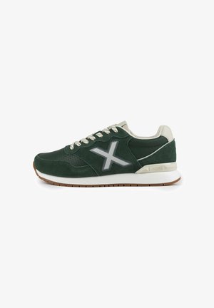 Zapatillas de ante verde con acentos beige, que presentan un logotipo plateado reflectante, detalles perforados y una suela blanca con una banda de rodadura marrón texturizada.