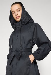 Black hooded jacket with a textured finish, cinched waist, and drawstring details. Features large pockets and a belted design.