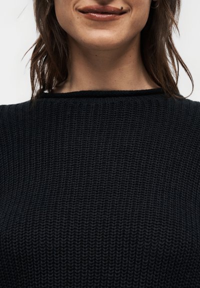 Γυναίκα με υφασμάτινο μαύρο πουλόβερ, κυματιστά καστανά μαλλιά, διακριτικό χαμόγελο και ελιά πάνω από το άνω χείλος.