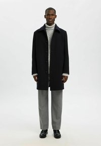 Vīrietis, kurš ģērbjas melnā pogātajā mētelī, pelēkajā turtleneck džemperī, pelēkās biksēs un melnās kurpēs, stāv uz vienkārša baltā fona.