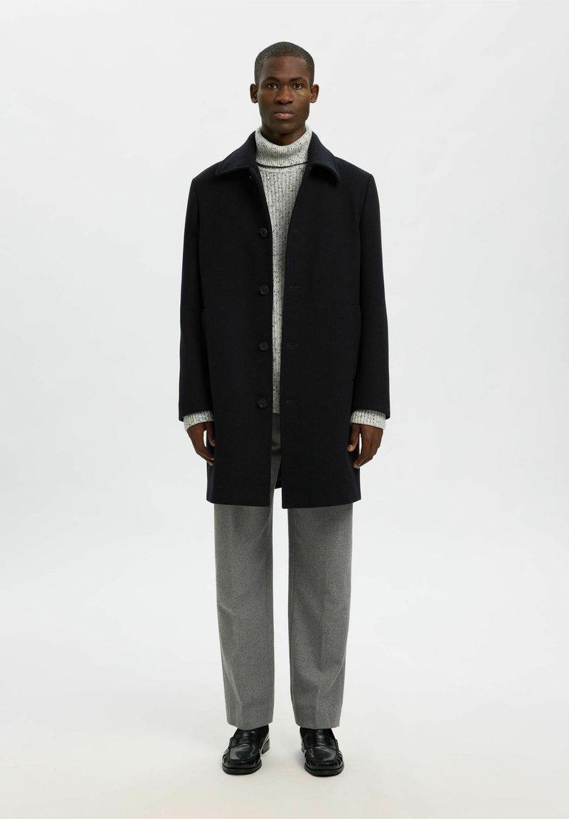 Vīrietis, kurš ģērbjas melnā pogātajā mētelī, pelēkajā turtleneck džemperī, pelēkās biksēs un melnās kurpēs, stāv uz vienkārša baltā fona.