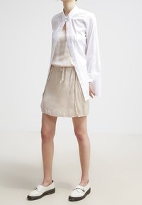 Chemise blanche boutonnée à manches longues portée sur une robe nuisette beige, avec une taille à cordon de serrage et une texture lisse. Chaussures noires et blanches.