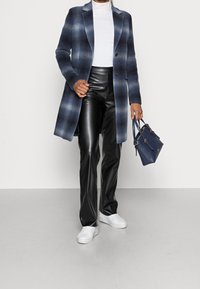Lange blauw en grijs geruite jas over een witte top, gecombineerd met zwarte leren broek en witte sneakers; houd een kleine blauwe handtas vast.