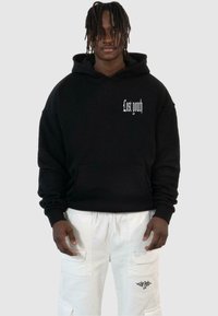 Black hooded sweatshirt with "Lost youth" printed in white, featuring a kangaroo pocket and loose fit. Worn with white cargo pants.
