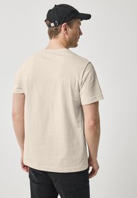 Beige bavlněné tričko s krátkými rukávy, rovného střihu a kulatým výstřihem. Nosené s černými kalhotami a černou baseballovou čepicí.