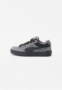 Zapatilla Puma en gris y negro con cordones negros, ojales naranja y una suela gruesa negra, mostrada en vista de perfil lateral.