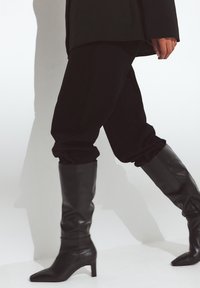 Botas de cuero negras hasta la rodilla con punta afilada y tacón afinado. Presentan una textura arrugada y un acabado suave.