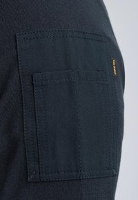 Dark blue fabric with a smooth texture features a patch pocket with double stitching and a yellow accent label on the side.