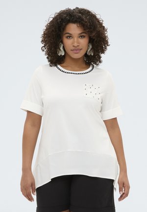 Kvinde med krøllet hår iført hvid kortærmet top med sortkantet krave og perlebroderet lomme, sat sammen med sorte shorts.