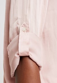 Ljusrosa skjorta med uppvikta ärmar och knappstängning, tillverkad av ett slätt tyg med en subtil glans och lätta vertikala ränder.