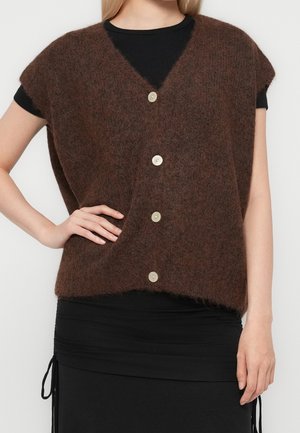Personne portant un gilet sans manches marron duveteux boutonné par-dessus un haut noir et une jupe noire avec des liens sur les côtés, main sur la hanche.