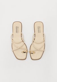 Beige veganske sandaler med en flad firkantet tå, der har buede anklestraps og gyldne ovale detaljer. Glat tekstur og minimalistisk design.