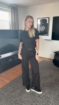 Oseba z dolgimi svetlo rdečimi lasmi, oblečena v črno majico, široke črne hlače in črne superge, stoji na sivem preprogi v dnevni sobi.