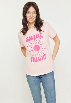 Vaaleanpunainen puuvillainen t-paita lyhyillä hihoilla, jossa on suuri vaaleanpunainen auringon grafiikka ja teksti "SHINE BRIGHT" paksuilla vaaleanpunaisilla kirjaimilla.